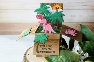 Die D-Lites Inspiration | Dinosaur Pop Up Card. Video tutorial by Nichol Spohr for Spellbinders using S2-273 Sun and Clouds, S3-317 Dinosaurs, S3-320 Picket Fence, S3-249 Palm Trees, S4-788 Classic A2 Waves Borders, S5-233 Heart & Home Scalloped Pop Up Box #spellbinders #popupbox #diecutting handmadecard #popupcard